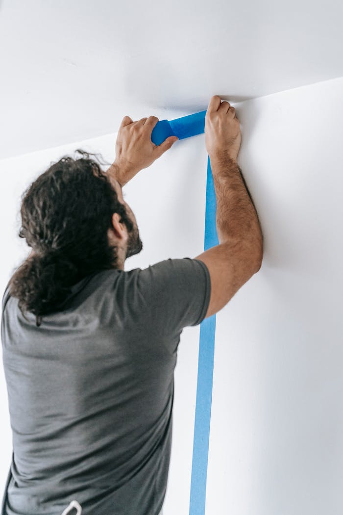 A man applying blue tape on a wall during a home renovation project, showcasing painting preparation.